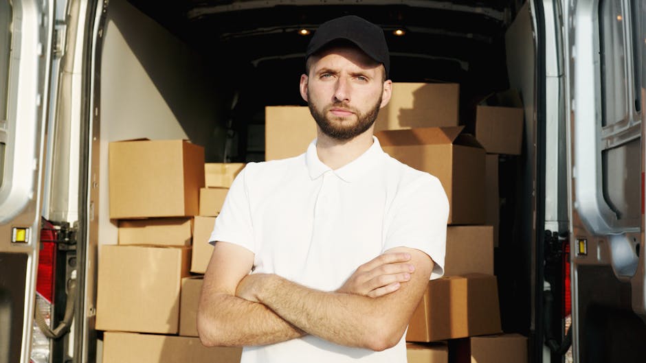 A man wearing a blue beanie, red and navy jacket, and black trousers stands confidently in front of the open rear doors of a van, which is loaded with stacked cardboard boxes for a home relocation. Th