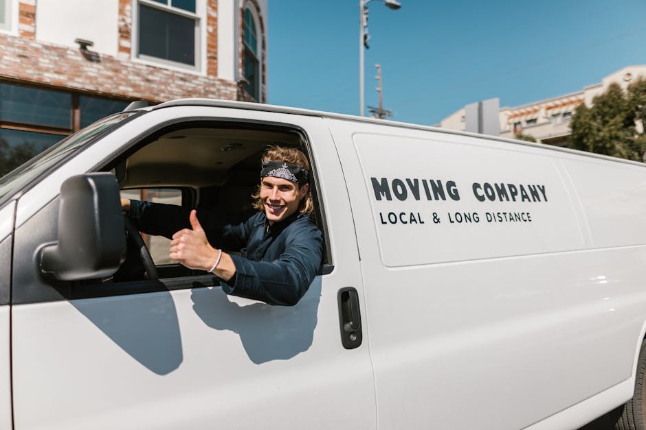 A smiling young man with long hair, wearing a black headband and a dark jacket, is sitting in the driver's seat of a white man with Van Harringay van. He is giving a thumbs-up gesture and looking towa