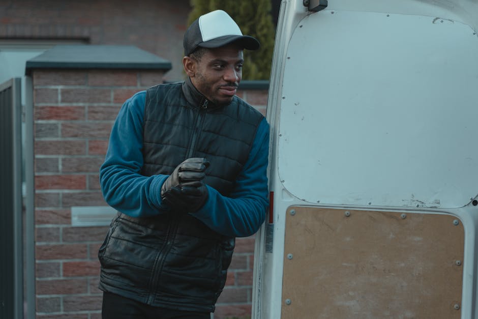 A man dressed in a blue long-sleeve shirt, black quilted vest, and wearing a black and white cap, stands next to a white delivery van during a home relocation or moving process. He is wearing black gloves and appears to be preparing to load or unload items. The van's side door is open, revealing the interior, which may contain packing materials such as cardboard boxes and possibly furniture or appliances. Behind him, a brick house and a garden are visible, indicating an outdoor setting on a residential street. The scene is well-lit with natural daylight, capturing a moment of furniture transport or packing and moving activities associated with professional removals, as offered by [COMPANY_NAME]. This image reflects standard equipment and procedures used in house removals and moving services in Harringay, supporting the logistics of home relocations.