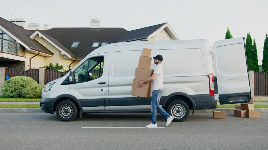 A man dressed in a blue long-sleeve shirt, black quilted vest, and wearing a black and white cap, stands next to a white delivery van during a home relocation or moving process. He is wearing black gloves and appears to be preparing to load or unload items. The van's side door is open, revealing the interior, which may contain packing materials such as cardboard boxes and possibly furniture or appliances. Behind him, a brick house and a garden are visible, indicating an outdoor setting on a residential street. The scene is well-lit with natural daylight, capturing a moment of furniture transport or packing and moving activities associated with professional removals, as offered by [COMPANY_NAME]. This image reflects standard equipment and procedures used in house removals and moving services in Harringay, supporting the logistics of home relocations.