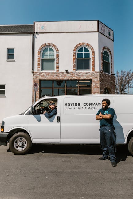 A white moving van parked on a street in front of a modern, two-storey building with a brick and white exterior, featuring arched windows on the upper floor. Inside the van's open side door, a man is visible, interacting with the driver who is wearing glasses and a dark jacket. Outside the van, a man with dark skin, wearing a blue shirt and jeans, stands with arms crossed, observing the scene. The van has the text 'MOVING COMPANY LOCAL & LONG DISTANCE' printed on its side, indicating professional removal and transport services. The environment appears bright with clear weather, and the scene conveys the early stages of a home relocation process, involving furniture and boxes inside the house or being prepared for loading.