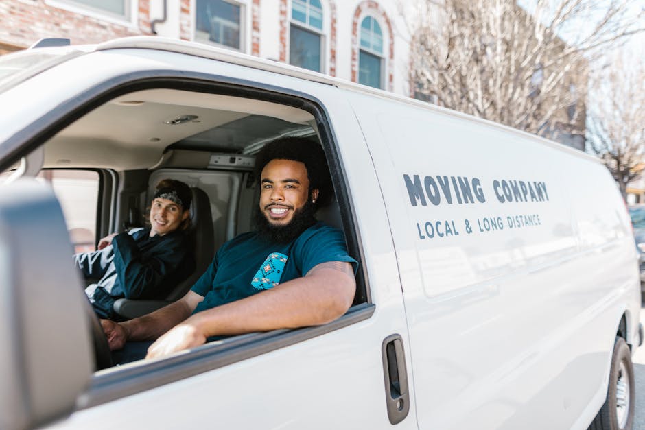 A man wearing a white t-shirt, blue jeans, and a black cap is engaged in a home relocation process, carrying three cardboard boxes of varying sizes outside a white van parked on a suburban street. The